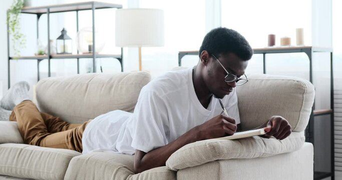 Young man lying on sofa and writing to do list in notepad at home