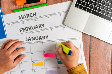 Top view of person using markers to highlight activities on calendar for the first few months of the year, next to his laptop. Several colored adhesive note. Concept of organizing tasks and objectives