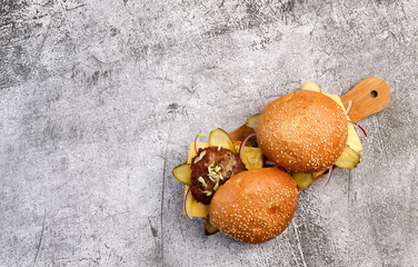 Homemade burgers with cheese, pickled cucumbers and pink onions on a small cutting wooden board on a dark grey background. Top view, flat lay.