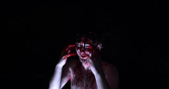 A maniac is covered in blood having a one on one psychotic episode in a darkened space with one single light placed at his feet