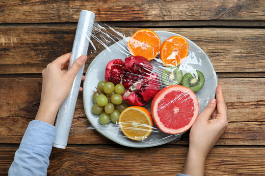 Woman putting plastic food wrap over plate of fresh fruits at wooden table, top view