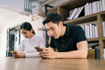 Young Asian male and female working together on the wooden table with laptop, looking at their mobile phones with a bookshelf in the background.