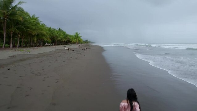 Aerial rising over young brunette woman walking near dense jungle in Dominicalito Beach shore on a cloudy day, Costa Rica