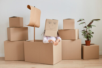 Unknown girl sitting surrounded with packages with personal pile, making head from cardboard box with drown smiling face, relocating, moving, real estate.