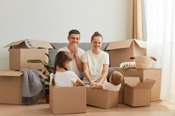Positive smiling young couple sitting on the floor near sofa surrounded with packages, looking at their children with happy smile, relocating, renting or buying new home.