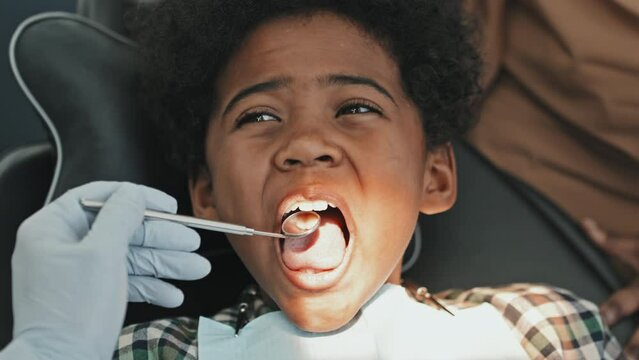 Close-up of cute elementary-aged Black boy sitting in dentist chair, opening mouth wide for unrecognizable doctor with hand in glove in frame only using dental mirror