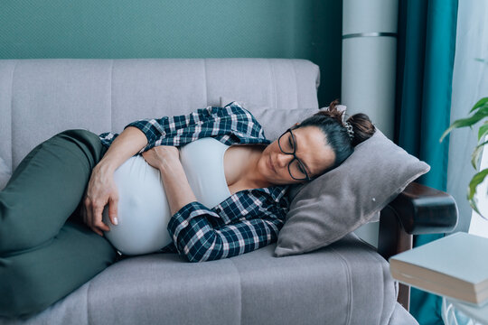 Dark-haired pregnant woman in and plaid shirt lying on comfortable grey couch in modern living room. Female feel pain or discomfort and hug belly. Contractions, braxton hicks 
