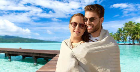 leisure, travel and tourism concept - happy couple in sunglasses covered with blanket hugging over...