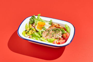 American cobb salad with chicken, eggs and vegetables in minimal style. Healthy salad on trend red background with hard shadow. Bowl with salad, chicken, eggs, vegetables in contemporary style