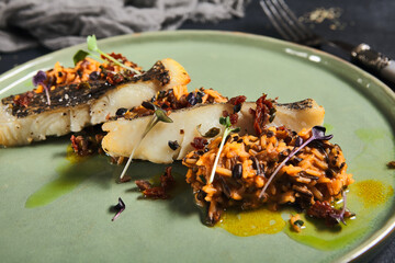 Risotto with slice roasted white fish in green plate on black background. Grilled cod with mix...