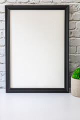 Black vertical frame beside a small houseplant, painting or artwork display mockup in front of vintage white brick wall.
