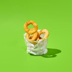 Fried onion rings in paper bag on green background. Flying onion rings in minimal style. Fast food appetizer in contemporary concept. Junk food on color background. Fast food menu.