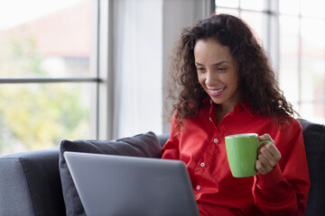 Happy smiling latin woman drinking coffee and using computer in living room. Lifestyle concept.
