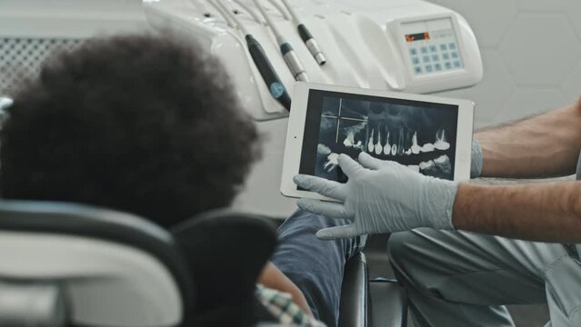 Over-shoulder of child patient sitting in dentist chair in modern clinic, looking at tablet computer with image of dental X-ray in hands of unrecognizable doctor at daytime