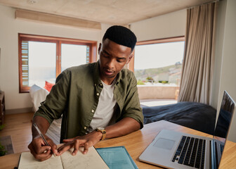 Young adult black male writing note in notebook next to laptop, concentrating. Sitting in modern...