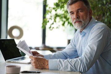 Fototapeta premium Serious mature male employee sits at the table in modern office, works with documents