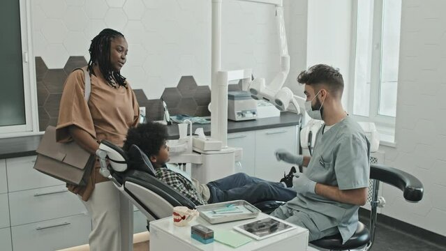 Locked-down of male Caucasian doctor starting medical procedures on Black ten-year-old boy who sitting in dentist chair, mother leaving modern medical office at daytime