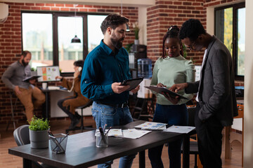 Fototapeta premium Diverse businesspeople analyzing marketing statistics on tablet computer working at company project in startup office. Multiethnic team brainstorming ideas planning business collaboration.