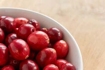 A white bowl with red ripe cranberries on a wooden background. Image with selective focus and copy space. 