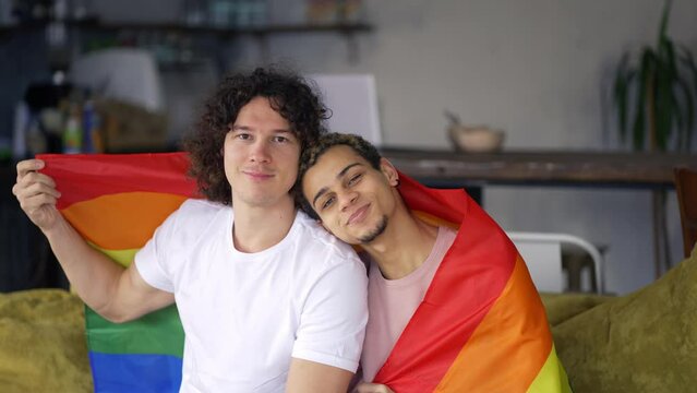 Guys spend time together at home on the couch, they hug and caress each other, under lgbt colorful rainbow flag