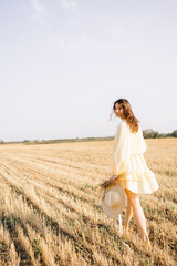 Naklejka na ściany i meble Caucasian young brunette walking through a wheat field. girl in yellow summer dress and straw hat. sheared golden ears of wheat, rye. Close-up portrait of a beautiful girl with brown eyes. 