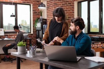 Businesswoman showing marketing charts to man leader discussing company strategy in brick wall startup office. Diverse businesspeople brainstorming ideas working at professional business collaboration