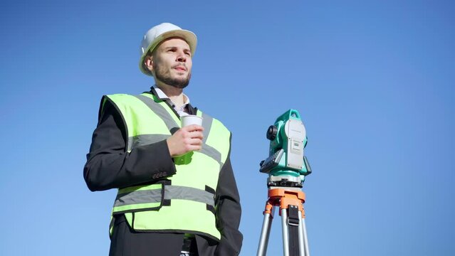 Medium shot portrait of confident engineer standing at background of clear blue sky drinking coffee. Handsome Caucasian man in hard hat and vest outdoors with theodolite. Lifestyle and occupation