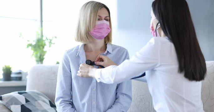 Doctor in protective mask listening with stethoscope to heart and lungs of female patient at home 4k movie