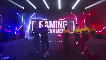 Group of teammates esport gamers walking and clapping hands then standing in front of monitor with gaming tournament banner at start of championship.