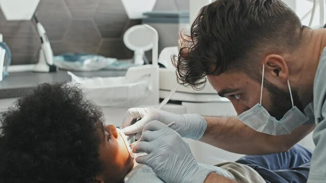 Tracking out of young male Caucasian doctor wearing mask and gloves, taking tool and starting treatment of teeth of Black boy lying in dentist chair in bright clinic