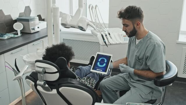 Medium long of bearded male Caucasian doctor wearing scrubs and gloves, showing image of perfect bite on screen of tablet computer to curly-haired boy sitting in dentist chair at daytime