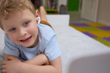 Close-up of a small blond boy in white wireless headphones using a modern laptop on the bed. The child watches entertainment content, listen to music on a portable device while lying on the bed