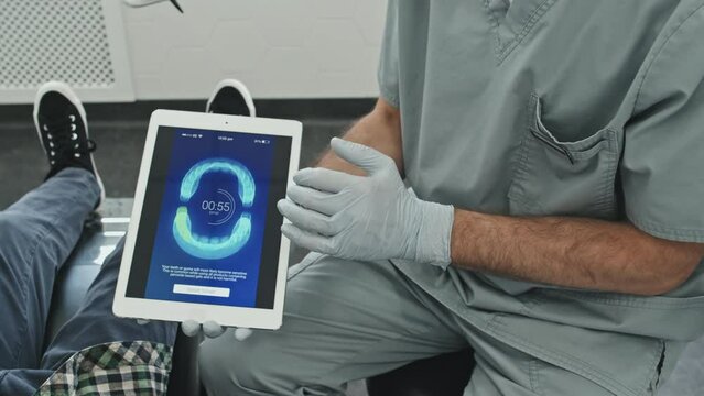 High-angle of cropped male doctor wearing scrubs and gloves, showing image of perfect bite on screen of tablet computer to curly-haired little patient sitting in dentist chair at daytime