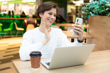 Young beautiful pretty brunette woman wearing white sweatshirt sitting at a shopping center at a table and working at a computer laptop, using cellphone. Freelance and business concept