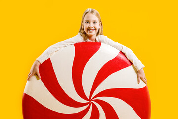 A cute satisfied girl holds a huge candy in her hands on a yellow background. Sweet gift. Large lollipop. Christmas sweetness.