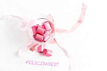 Chocolates with pink foil in a glass cup with pink bows on a white background and congratulatory letters top view