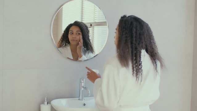 Beauty care at home. Young positive african american woman applying pampering cream on her face, looking at mirror