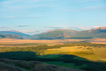 Scenic aerial view to mountain village near forest and sunlit golden steppe against high mountain...