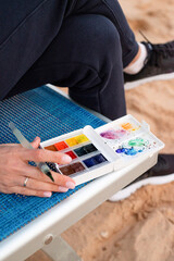 Closeup of watercolor palette, young woman artist painting or doing travel sketches using watercolor by the seaside