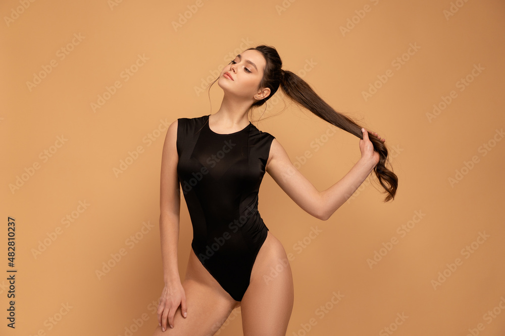 Wall mural Young beautiful woman dressed seductive black bodysuit standing studio isolated on color background holding hair ponytail looking away