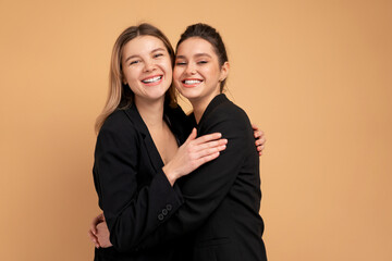 Two young business woman dressed black suit laugh and happy. Two beautiful caucasian female business person hugging isolated on beige color background