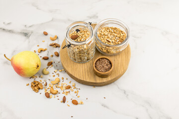 two different glass jars with walnut granola stand on a cutting round board. a marble background with scattered nuts on it and a ripe pear.