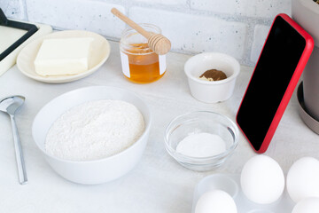 Fototapeta na wymiar The concept of online lessons in the preparation of cookies. A smartphone in a red case and ingredients are on the table. Everything is ready for mixing future baking. Horizontal Orientation