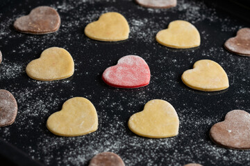 Macro view of pre-baked vanilla, pink and chocolate cookies in shape of hearts dusted with flour on black baking sheet