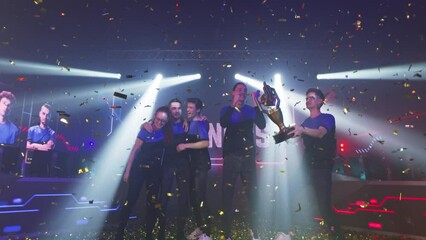Professional cybersport team of gamers winners with golden trophy hugging and jumping amidst confetti and pyrotechnics while celebrating victory in esports competition