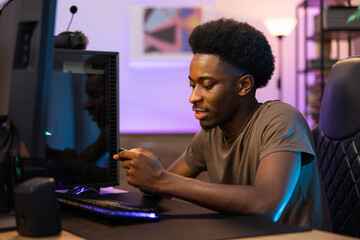 Portrait of dark-skinned man sitting comfortably in chair at gamer's desk boy is holding phone and...