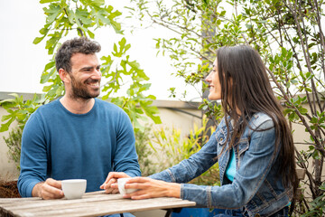 Young pouple in love drinking hot drink outdoor in a terrace, happy couple talking anf flirting, sitting on a table of a coffee shop with a cup of coffee or tea, leisure and relax lifestyle concept