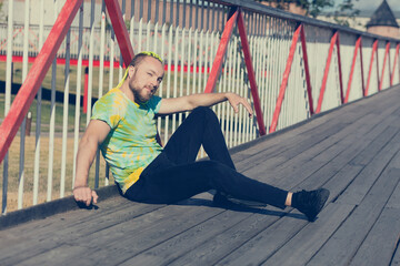 A man with an interesting mohawk hairstyle sits on the bridge and covers his eyes from the sun with...