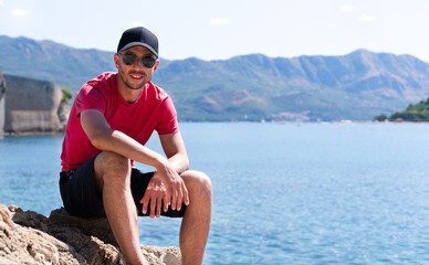 Smiling man tourist in a cap rests on the rocks on the seashore. Boka Kotorska bay in Montenegro. Copy space.