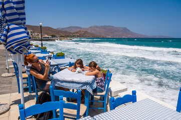 Family sitting seaside on blue chairs and table, typical Greek tavern in Kissamos, Crete, Greece